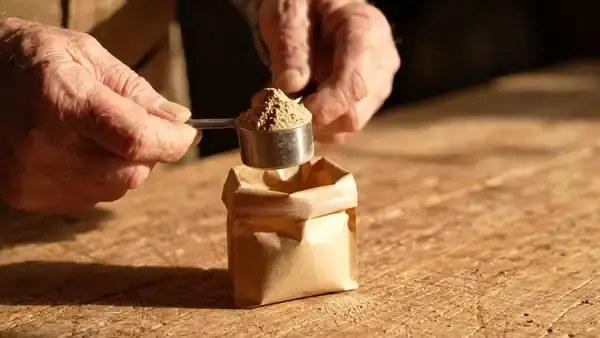 A man's hands levelling a heaped scoop of tan powder over an open pouch with a fingertip