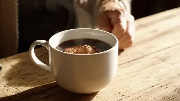 A teaspoon stirring tan powder into a white mug of black coffee until it disappears