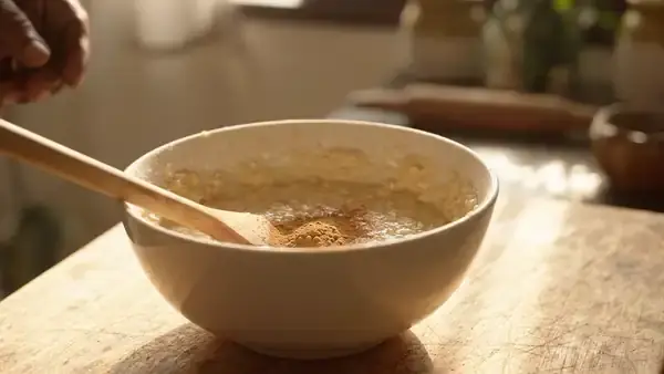 A man's hands folding tan powder into pale oat batter in a ceramic mixing bowl with a wooden spoon